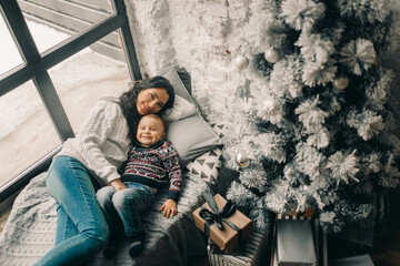 Happy mother and her son lies on windowsill near Christmas tree and gift box.