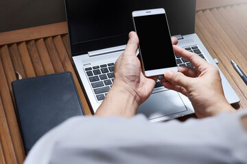 A cropped hand using a smartphone with a laptop and notebook on a wooden table