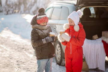 A girl and boy drink hot coffee, standing in the winter on the road next to a family car
