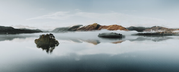 Misty morning on the lake with island reflections and low blue clouds. The Lake District, Cumbria. Autumn colours.  