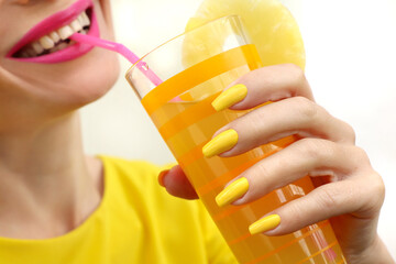 Young woman drinking pineapple juice with joy. Trendy manicure with yellow nail polish on a long...