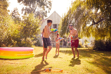 Family Wearing Swimming Costumes Having Water Fight With Water Pistols In Summer Garden