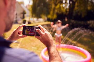 Father Taking Photo Of Daughter Playing In Paddling Pool On Mobile Phone