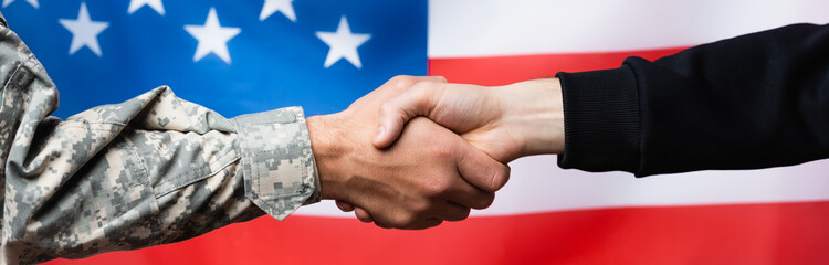 cropped view of soldier shaking hand with civilian man near american flag on blurred background,...