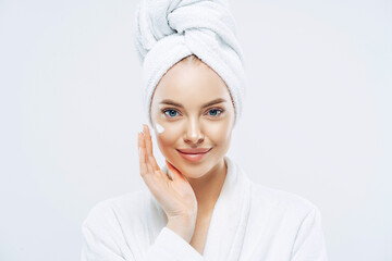 Young confident Caucasian woman applies face cream, enjoys new anti wrinkle cosmetic product, prevents sign of skin aging, wears minimal makeup, dressed in bath robe, isolated on white wall.