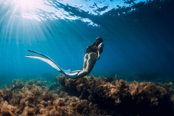 Freediver young woman with white fins glides undersea with sun rays. Freediving in transparent sea