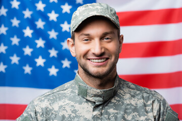 happy military man in uniform and cap smiling near american flag on blurred background