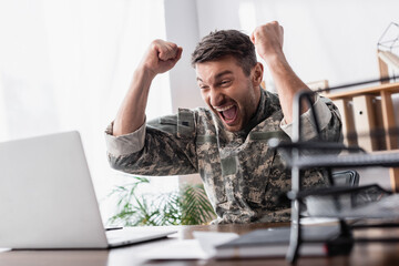 excited military man rejoicing near laptop and document tray on blurred foreground