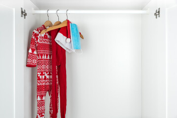 White and red christmas pajamas, hat, tights and face mask hanging on wooden hangers inside a white closet. Holidays during coronavirus pandemic..