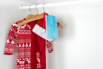 White and red christmas pajamas, hat, tights and face mask hanging on wooden hangers inside a white closet. Holidays during coronavirus pandemic..