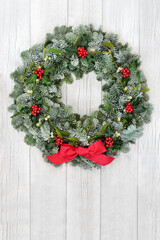 Christmas holly, mistletoe & snow covered spruce fi winter wreath with red bow on rustic wood front door background. Traditional theme for the solstice, xmas & New Year. Copy space.