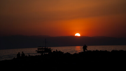 silhouette people and boat by the sea as the sun sets behind the hills