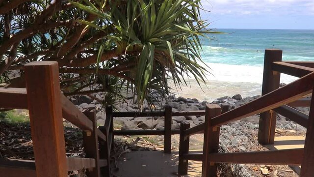 Timber Staircase Leading To The Turquoise Ocean With Surfers In The Distance Catching Waves And Lush Green Leaves