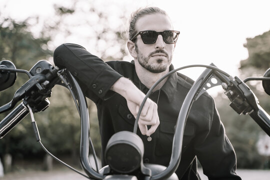 Unsaturated photo of a man with sunglasses leaning against the handlebars of a motorcycle outdoors