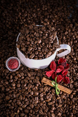 White cup full of coffee beans on a coffee beans background with red chocolate candy and red flowers. Morning espresso. Coffee mug. 