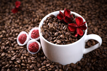 White cup full of coffee beans on a coffee beans background with red chocolate candy and red flowers. Morning espresso. Coffee mug. 