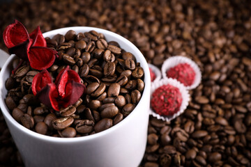 White cup full of coffee beans on a coffee beans background with red chocolate candy and red flowers. Morning espresso. Coffee mug. 