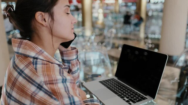 Caucasian attractive woman talking on the phone while working with laptop in cafe. A freelance girl communicates with someone.