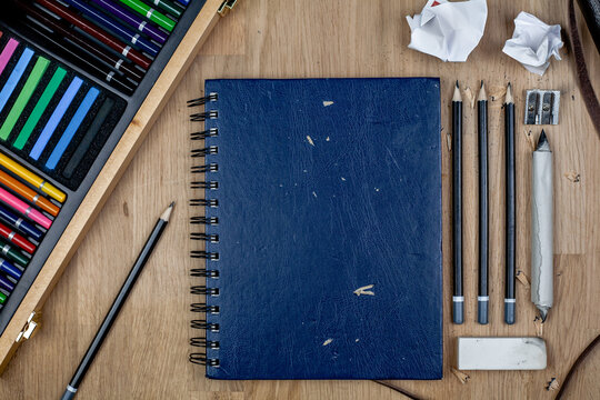 Blue sketchbook & stationary for sketching & drawing flat lay on a wooden background with colored pencils & pastels in a storage box on the left. Art organised creating background flat lay