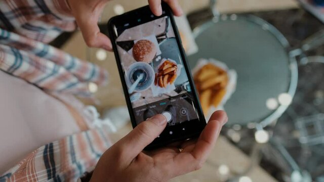 A woman uses her phone to take pictures from above of a hamburger, fries and cola. Tourist girl having a brunch or lunch at a fast food cafe.