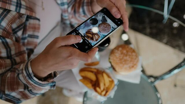 A woman uses her phone to take pictures from above of a hamburger, fries and cola. Tourist girl having a brunch or lunch at a fast food cafe.
