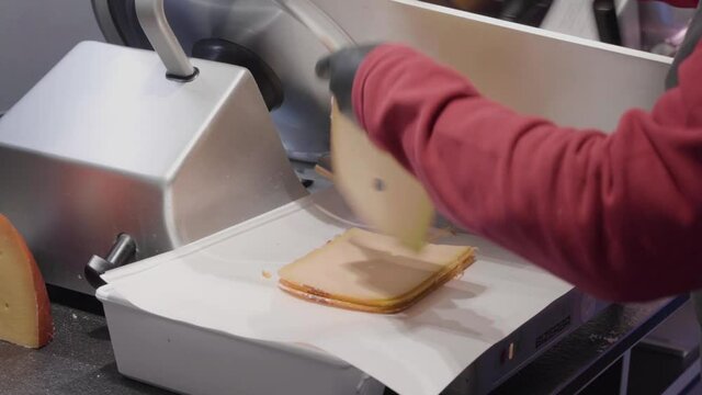 A uniformed, gloved deli worker slices cheese using an industrial cutter
