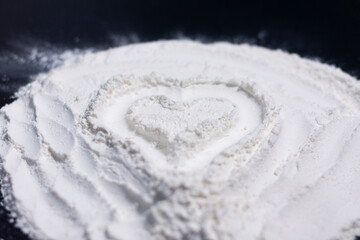A pile of flour on a dark surface. Drawing of a heart. Side view. 