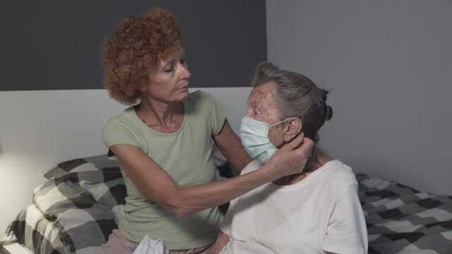 Mature caucasian nurse, volunteer assistant in nursing home help senior woman put on protective mask sit on bed. Danger contracting infection, virus old people during coronavirus epidemic covid 19