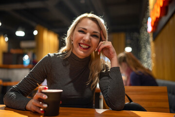 A blonde smiling girl is drinking coffee in a cafe in the evening.