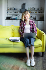 smiling blonde woman in checkered shirt working from home with laptop on knees