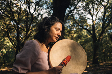 Fototapeta na wymiar young woman playing tambour in the woods in nature with incense. mystic woman doing ritual and ceremony . musician woman playing tribal drums