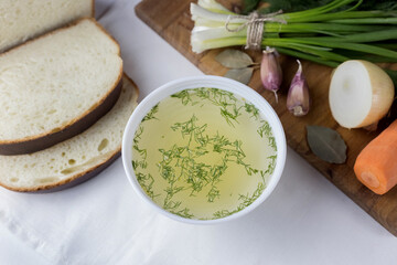 A cup of vegetable broth is on the table, next to it are vegetables and fresh herbs, bread, light photo