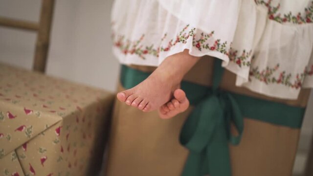 Slow-motion of Cute Girl dressed in festive dress sitting on Christmas Gifts swinging her feet - close up