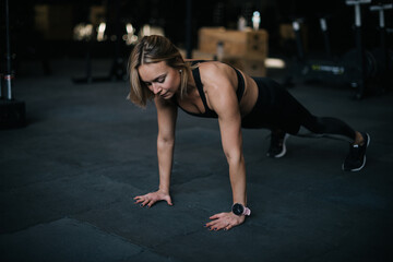 Fototapeta na wymiar Fitness young woman with perfect athletic body wearing black sportswear doing push-ups exercise on the floor with black mats during sport workout training at modern gym with dark interior.