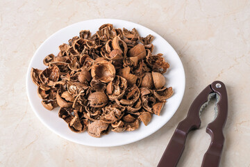Pile of empty broken walnut shells on a white plate and nutcracker over kitchen table. Biodegradable food waste and peelings. Nut crop concept.