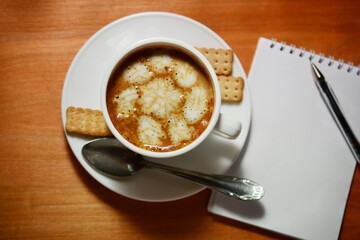 frothy coffee in a white cup on a wooden table. pen and notebook, concept of dating, rest, relaxation. creative plans.