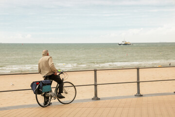 homme à vélo sur le bord de mer à Oostende en train de regarder un bateau au loin - obrazy, fototapety, plakaty