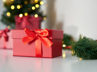 Close up of a red gift box. Beautiful gift box with a red ribbon and a bow. Background with green Christmas tree and lights. Christmas time and gift time