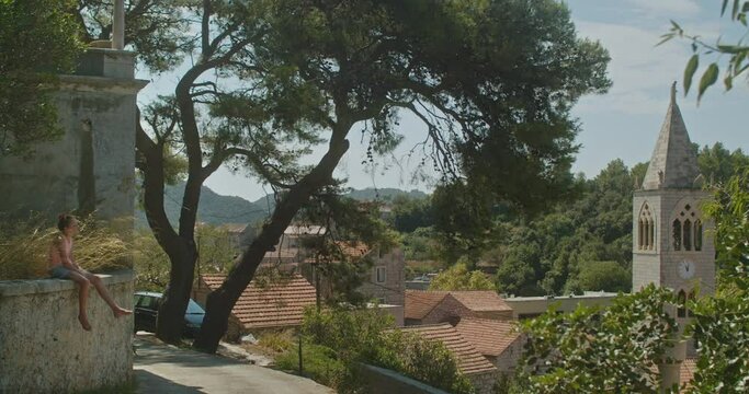 Cute teenage girl sitting on her own in the grass nearby a church in the middle of an emprty old town in Lastovo island, Croatia