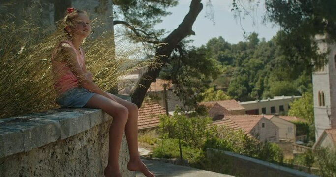 Cute teenage girl sitting on her own in the grass nearby a church in the middle of an emprty old town in Lastovo island, Croatia