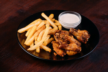 Fried chicken wings and potato fries with garlic sauce on black plate isolated on dark wooden background 