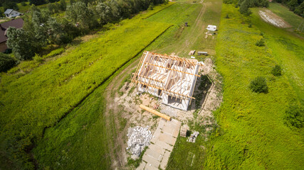 Aerial view on detached house under construction. House in basic state. Located on green plot in small village. Wooden roof structure. Meadows around.