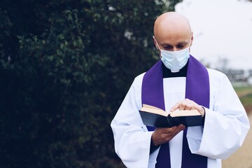 Priest in a medical mask reading holy bible.
