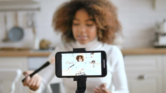 Kinky haired woman blogger with dark skin films makeup tutorial sitting against smartphone in modern studio closeup slow motion