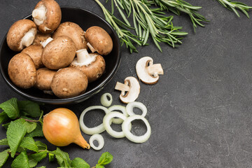 Obraz na płótnie Canvas Champignon mushrooms in a black bowl. Chopped onions and mushrooms, onion head on table. Sprigs of mint and rosemary
