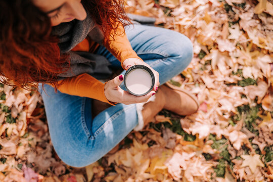 Top view and close up of a brunette woman sitting on the ground surrounded by leaves while holding a cup of coffee on her hands. Lifestyle autumnal outdoors.