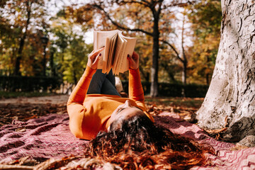 A pretty woman lying down on a blanket on the ground while reading a book. She is enjoying a break...