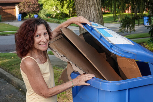 Pretty smiling woman senior citizen seen lifting the lid of a blue recycling can overflowing with cardboard boxes