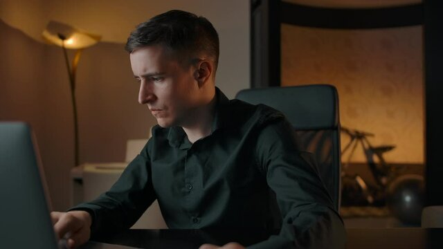 A young man works on a laptop, typing on a keyboard. Cozy apartment with evening light, refocusing from face to laptop screen