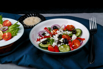 White plate with grilled zucchini with cherry tomatoes on lettuce and Greek salad on a blue napkin with a fork beside sesame seeds.